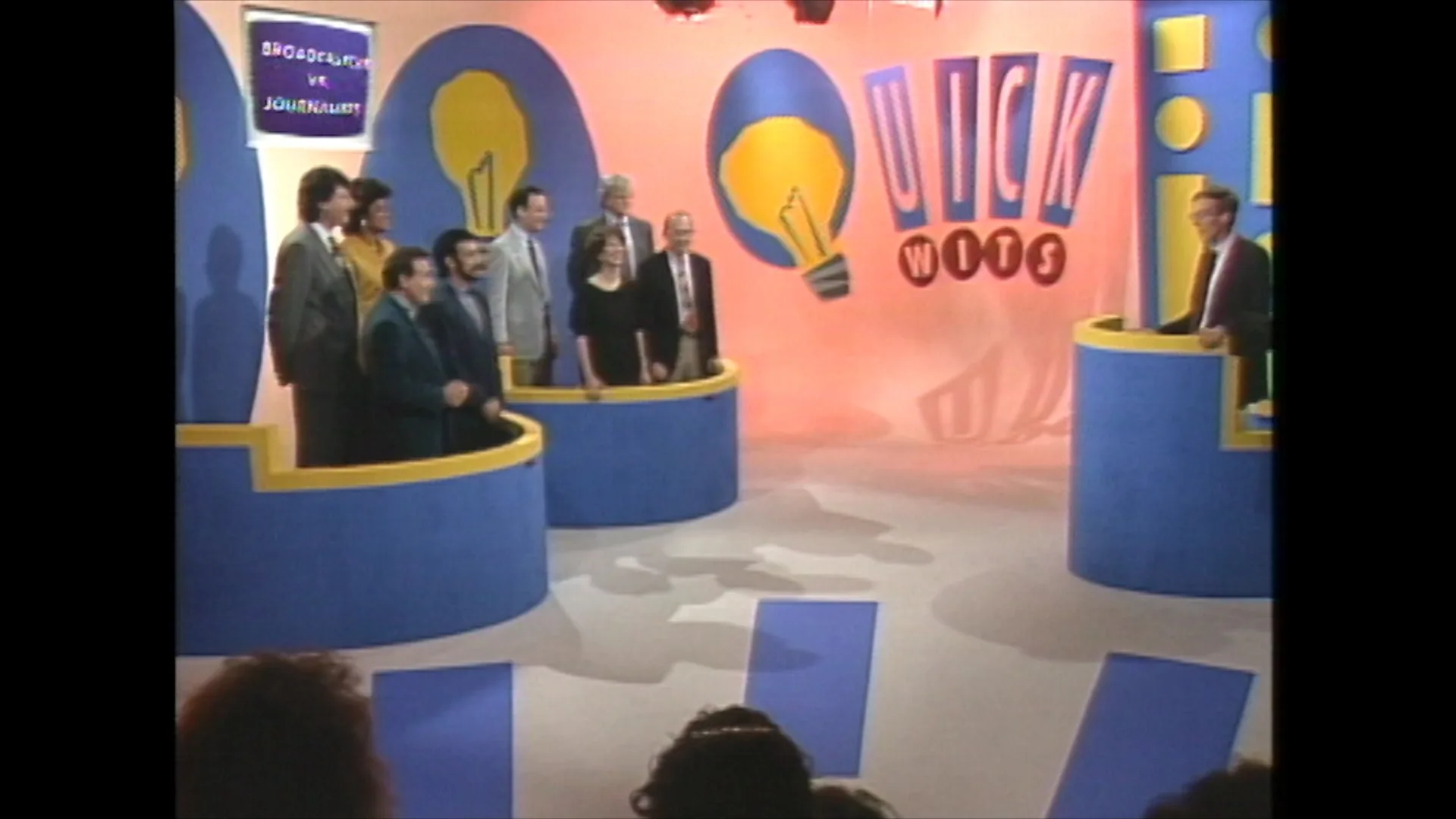 The colorful set of the "Quick Wits" TV game show with contestants lined up behind podiums