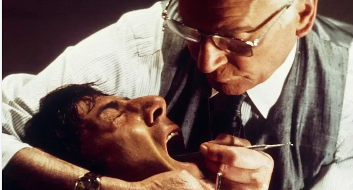 Film still of a menacing dentist leaning over a patient's open mouth, evoking dread of the drill-and-fill