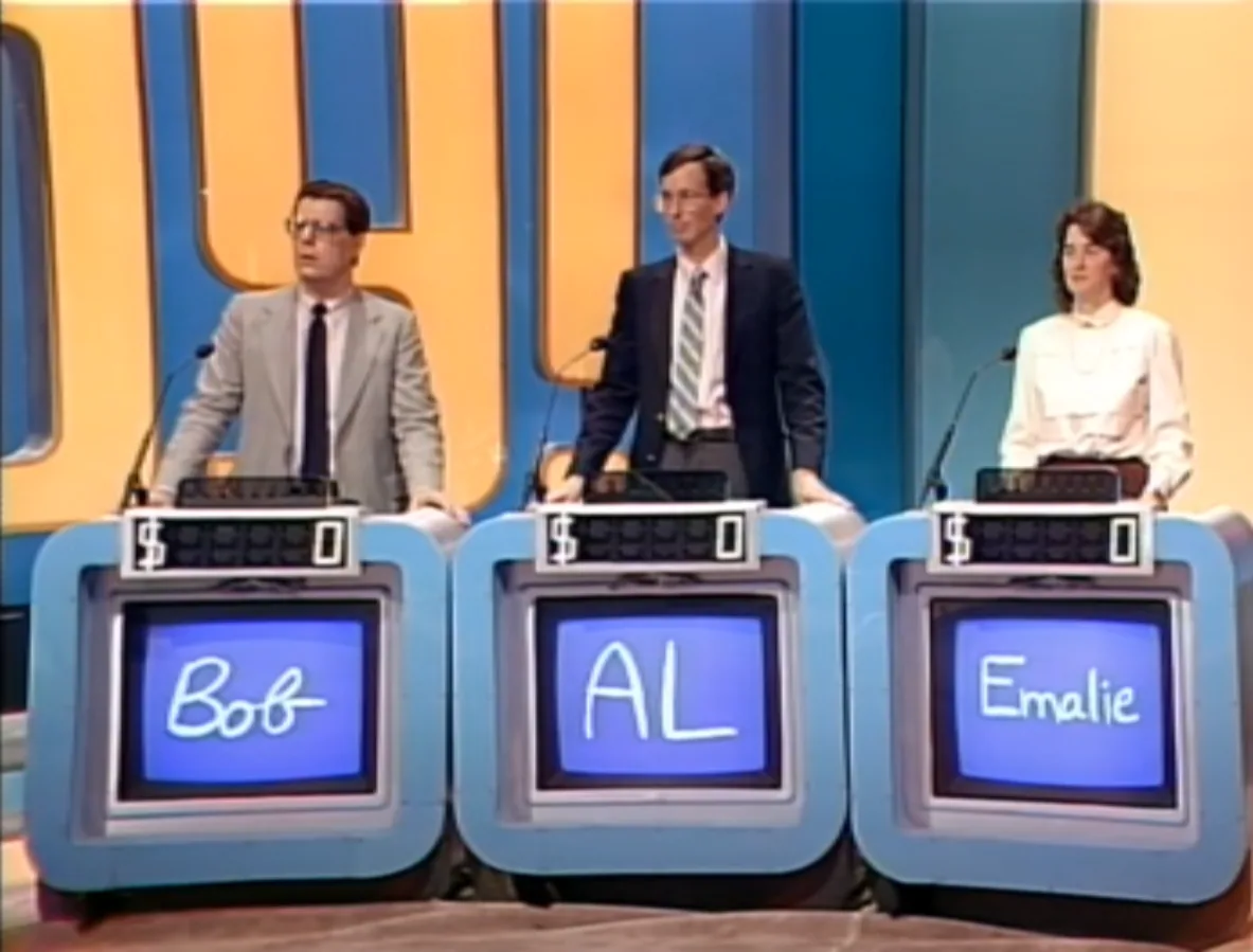 Al Lewis at the centre podium on the Jeopardy set, his nameplate reading “AL”, between two other contestants.