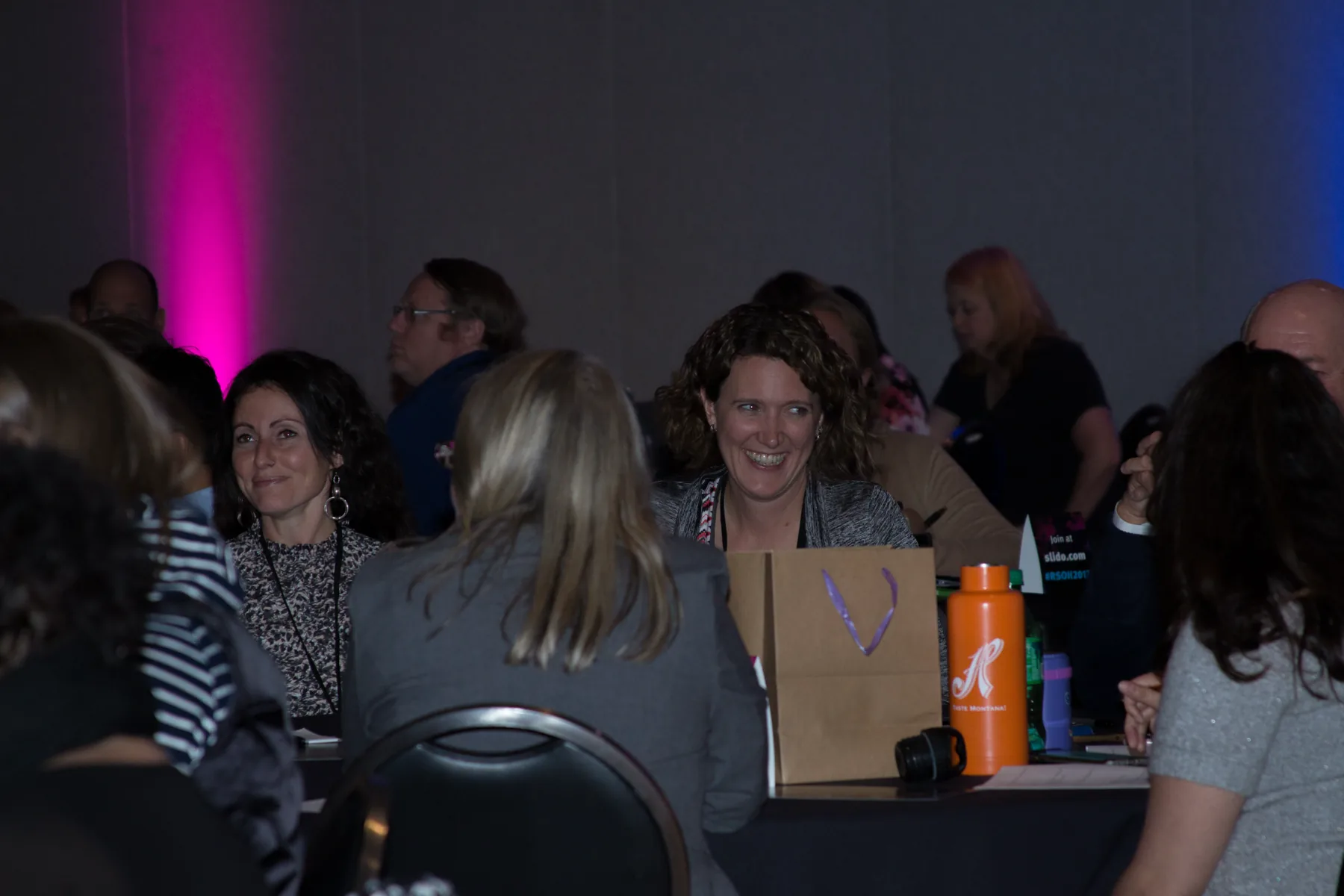Two attendees grinning across a table mid-contest, conference tote bags and water bottles in front of them.