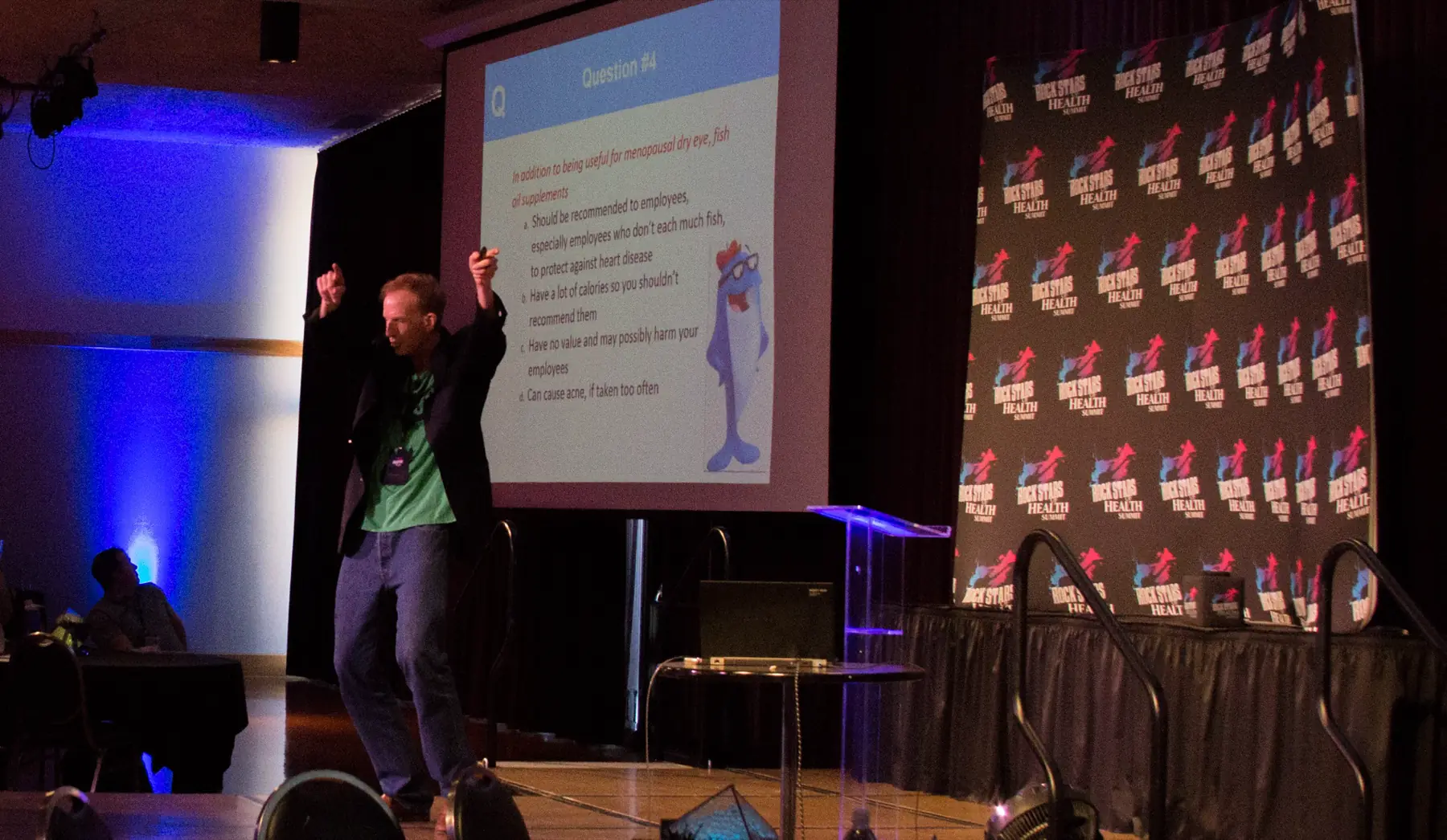 Al Lewis on stage with both arms raised, a Quizzify question projected on the screen behind him.