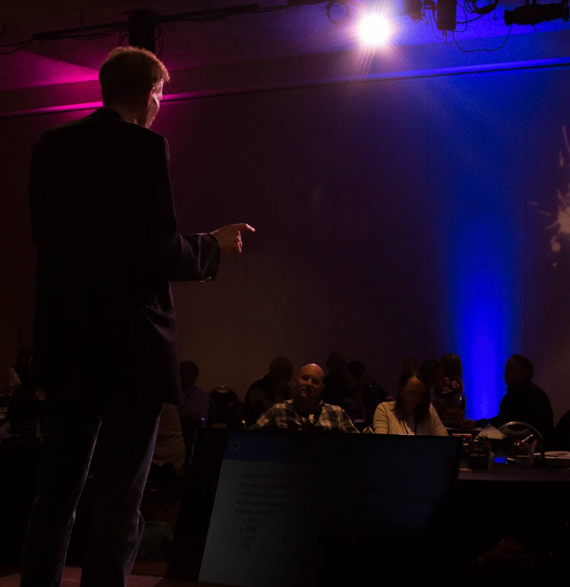 Al Lewis silhouetted against stage lighting, running a contest for a seated audience.