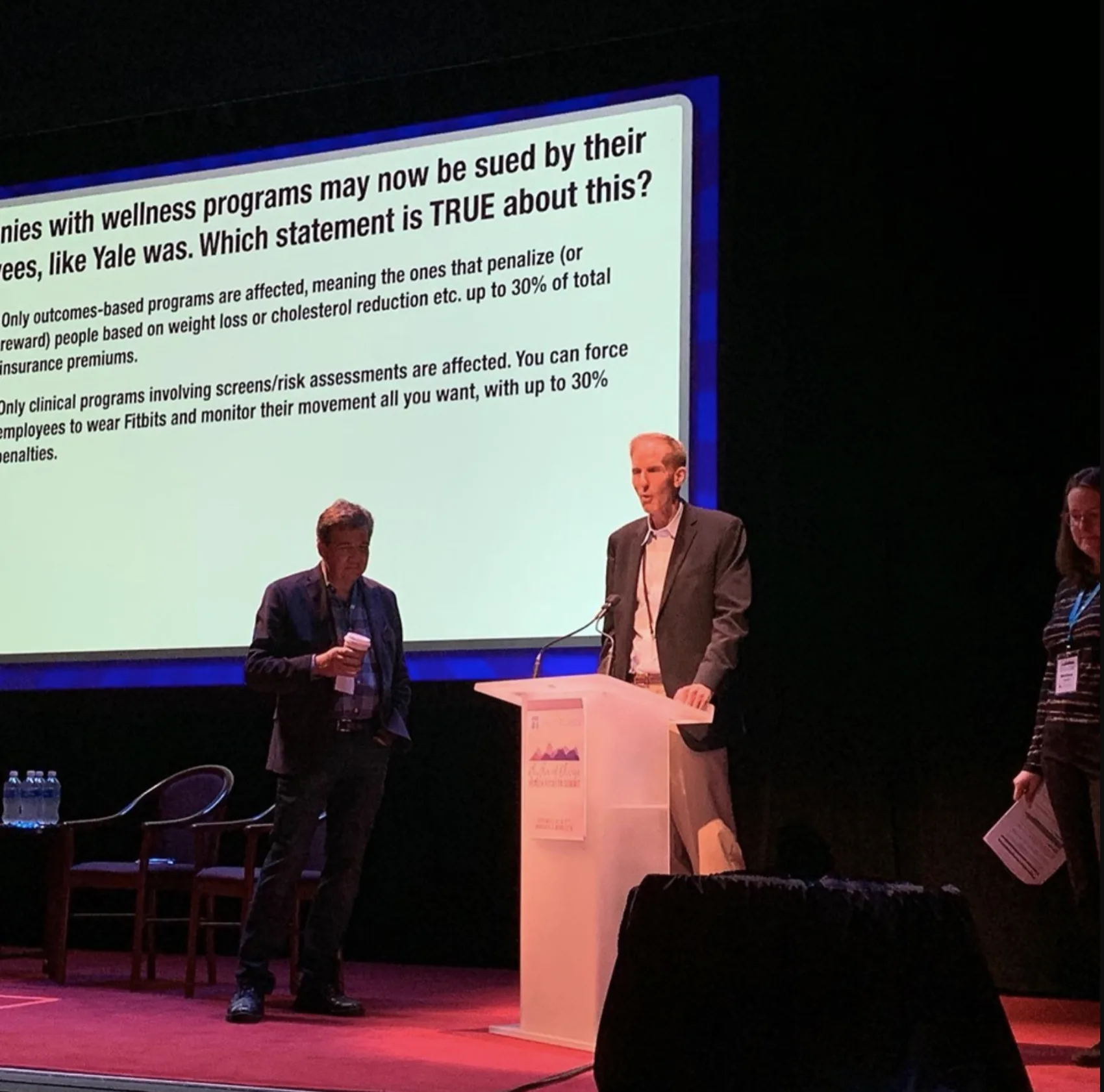 A Quizzify question on a ballroom screen asking which statement about wellness-program lawsuits is true, with two presenters on stage below it.